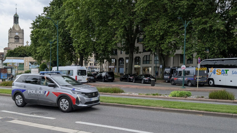 Véhicule de Police Nationale en circulation devant des fourgons et berlines officielles, bus Éva'D et clocher en arrière-plan